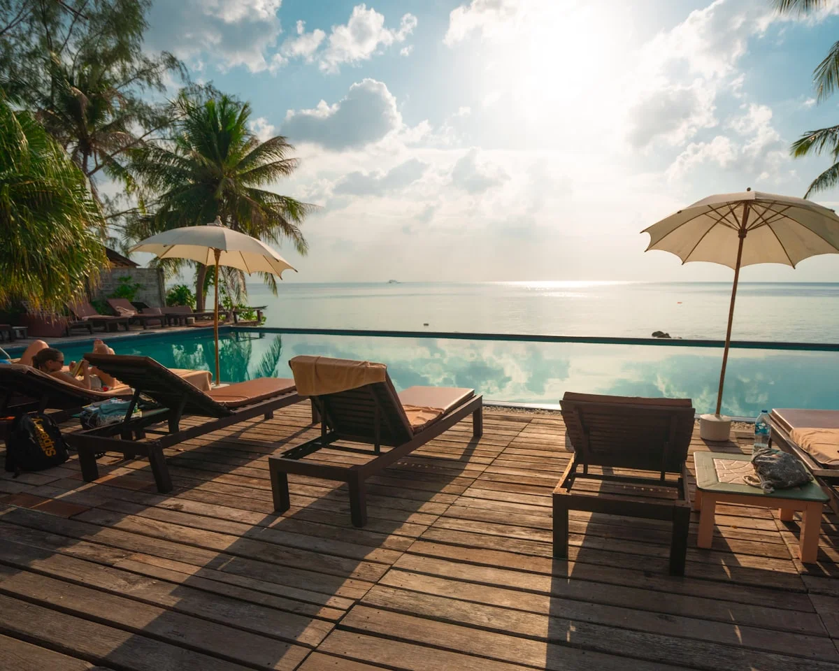 Timber pool deck with sun loungers and white parasols beside an infinity pool, facing a calm ocean and palm trees.